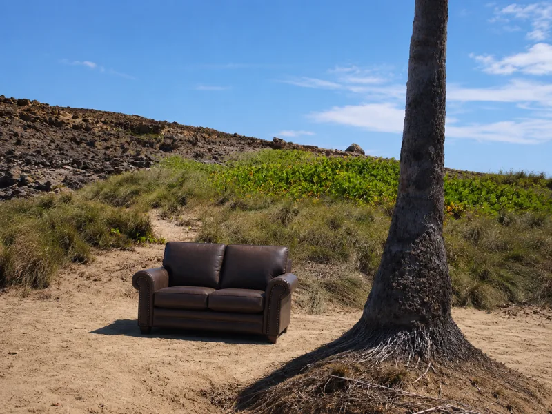 An abandoned couch on the sand beside a palm tree, under a bright blue sky
